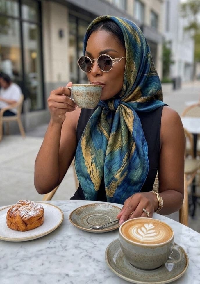 Woman wearing a blue patterned headscarf and sunglasses sips coffee at an outdoor cafe table.